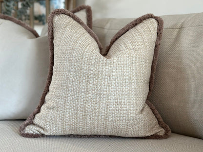 A handmade bespoke cushion with a textured woven fabric in a neutral tone, featuring a plush beige fringe trim. The cushion is placed on a beige sofa, adding a cosy and elegant touch to the decor.