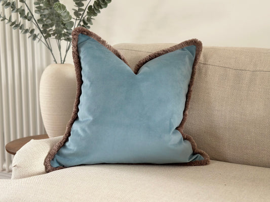 A sky blue velvet cushion with a beige taupe fringe trim Sitting on a sofa in an elegant costal decor scheme.