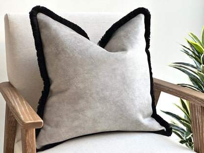 A light grey cushion with a subtle textured fabric and a black fringe trim.