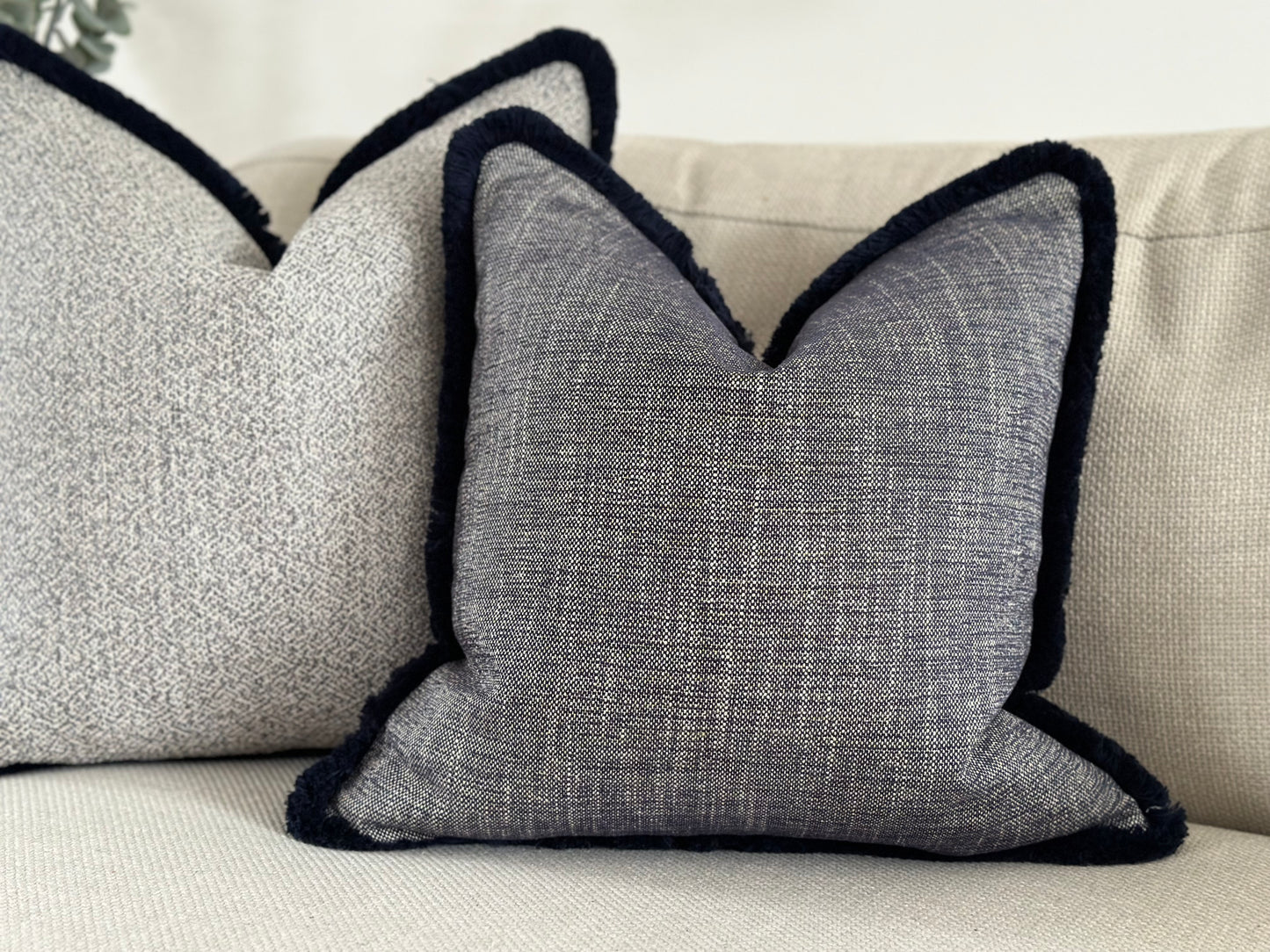 The best luxury pillow brand that is affordable. In the image is a beautiful luxurious textured navy blue cushion with a  fringe trim sitting in a sofa in a modern neutral livingroom also suitable for styling bed and chairs.