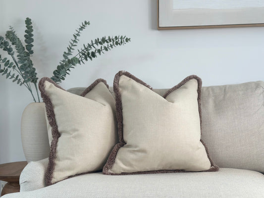 Two beige decorative pillows with brown plush fringed edges are placed on a light-colored sofa. The overall setting is minimalistic and cozy, with neutral tones dominating the decor.