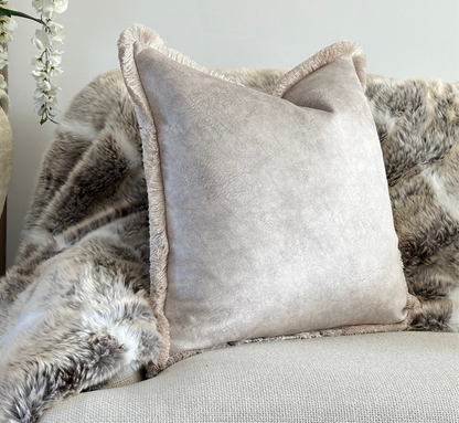 A luxurious square cushion in a soft, muted neutral ivory shade with a subtle texture and delicate fringe trim