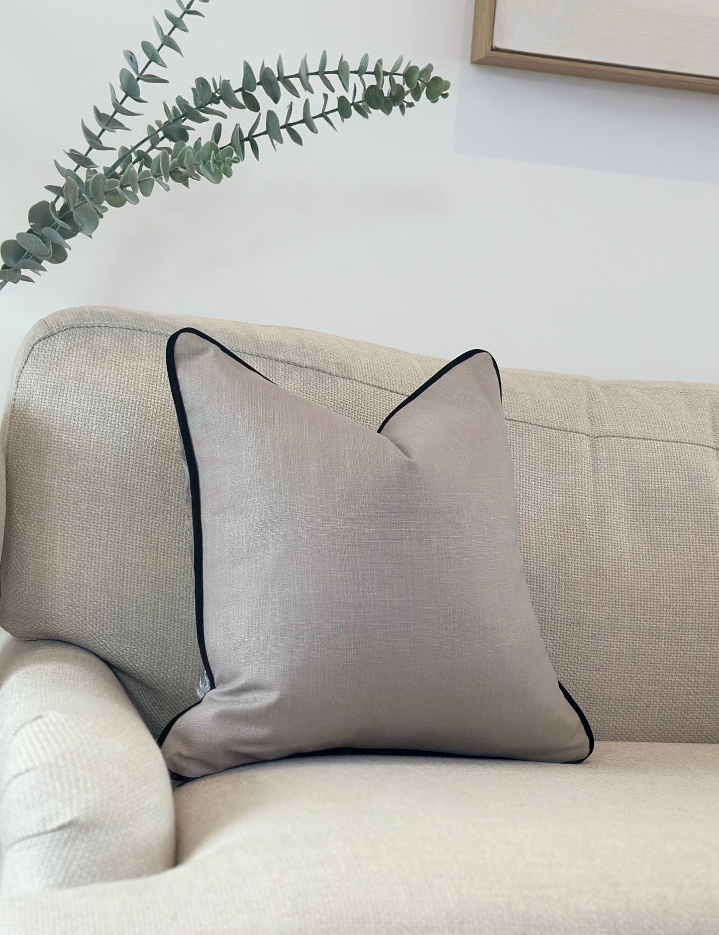 A Grey decorative pillow with a smooth, slightly textured fabric is placed on a light-colored sofa. The pillow is accented with a black piping around the edges, adding a subtle contrast adding a natural element to the minimalistic and serene setting.