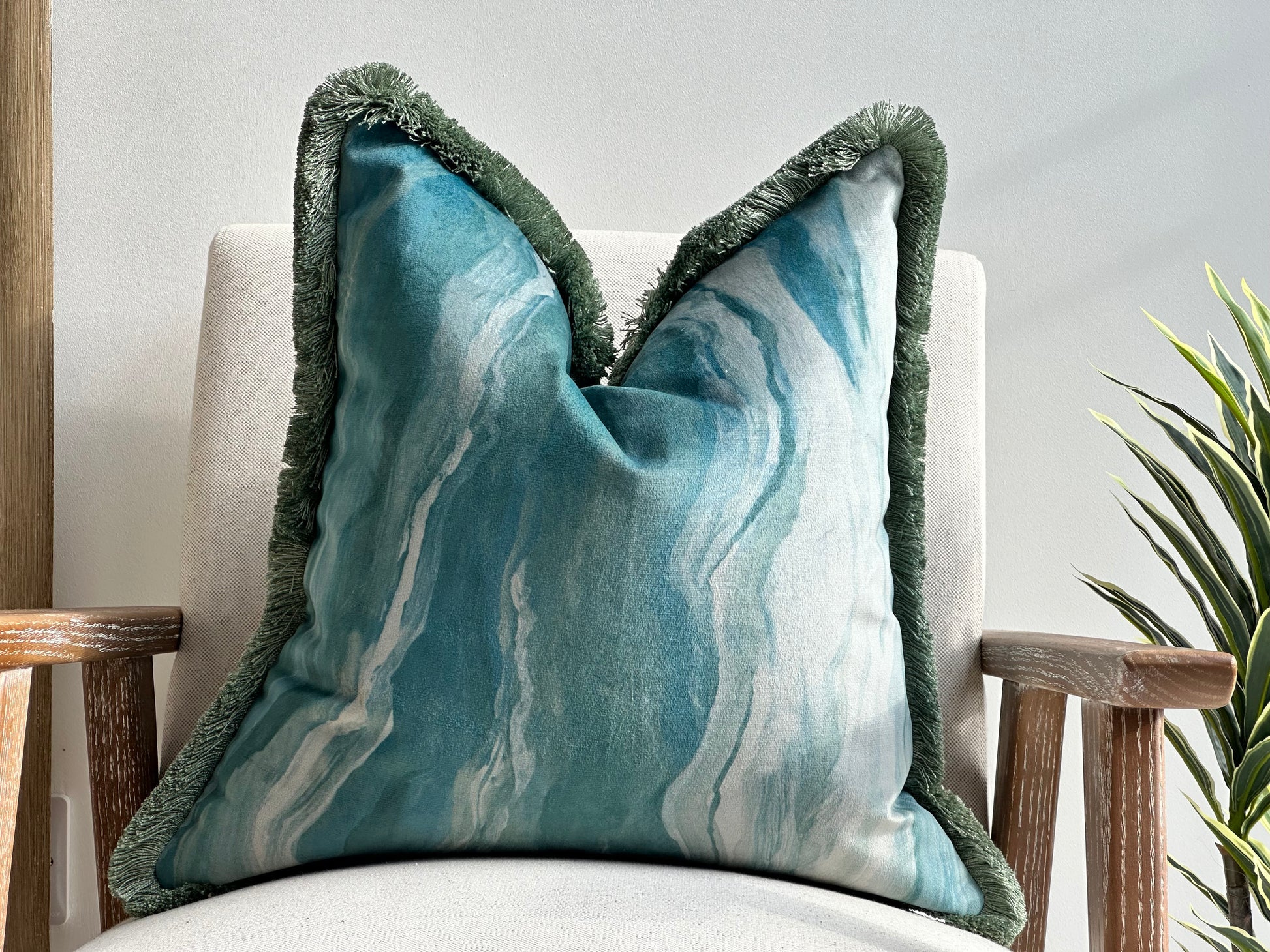 A square decorative cushion with a blue-green marbled fabric and an antique green fringed trim, placed on a neutral-toned armchair. The cushion features a luxurious, velvety texture and an organic, flowing pattern, complementing the cosy, calm costal setting.