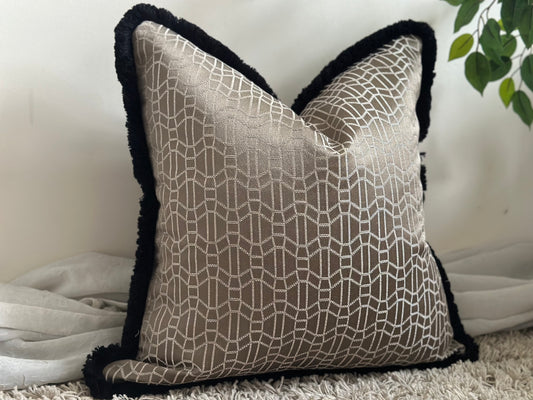 a decorative pillow. It has a beige or light tan fabric with a geometric pattern outlined in white, and a black, fluffy border.