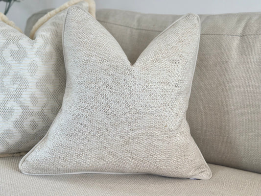 A light ivory beige throw pillow with a textured, woven fabric on a matching light-colored sofa. Behind it is another pillow with a similar colour scheme and a geometric pattern.